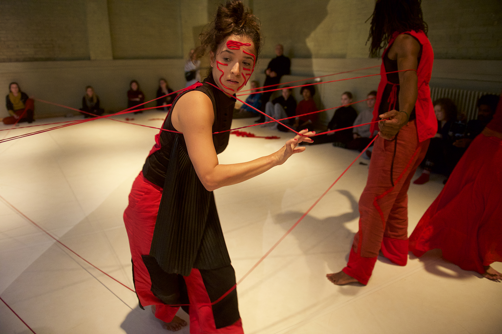 Dancers in black and red costumes, wrapped in a long red thread; and a doll made of fabric scraps and used clothes.