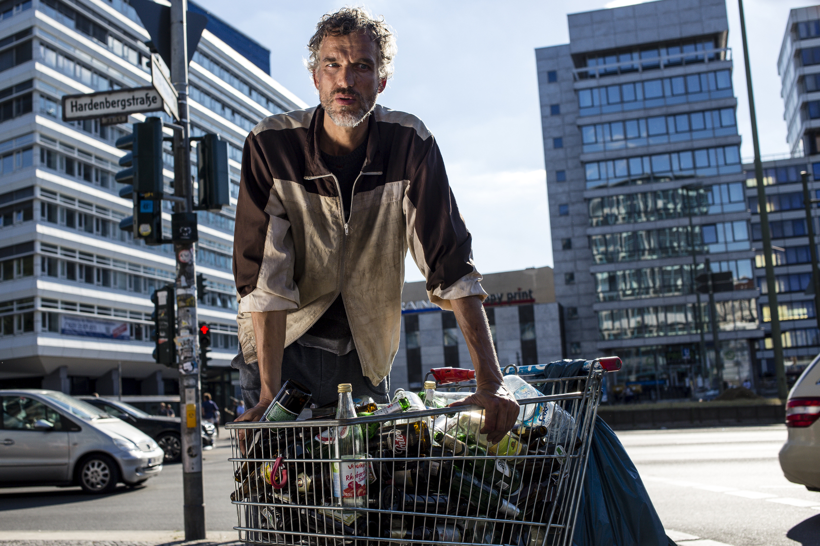 Man in grimy clothes leans on a supermarket cart full of empty bottles, he is on a big city street.