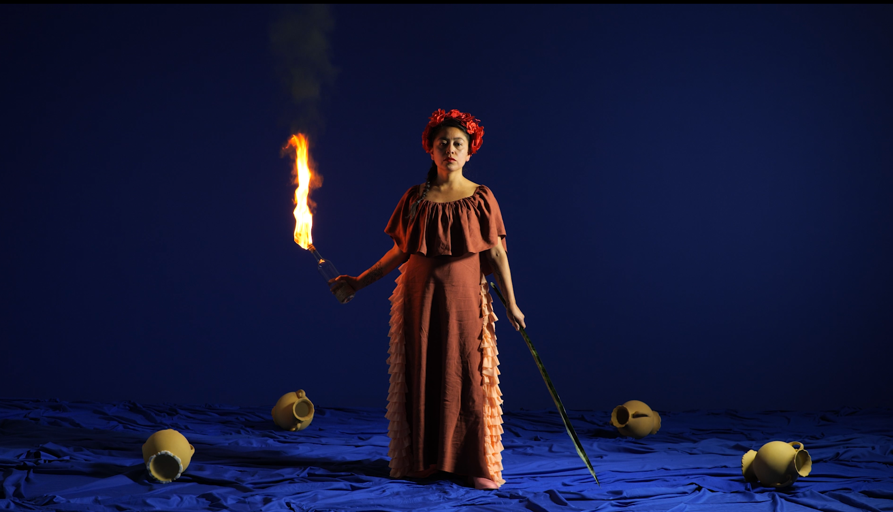 The performers are on a set with a royal blue background, one wearing costumes in shades of pink and the other in terracotta tones.