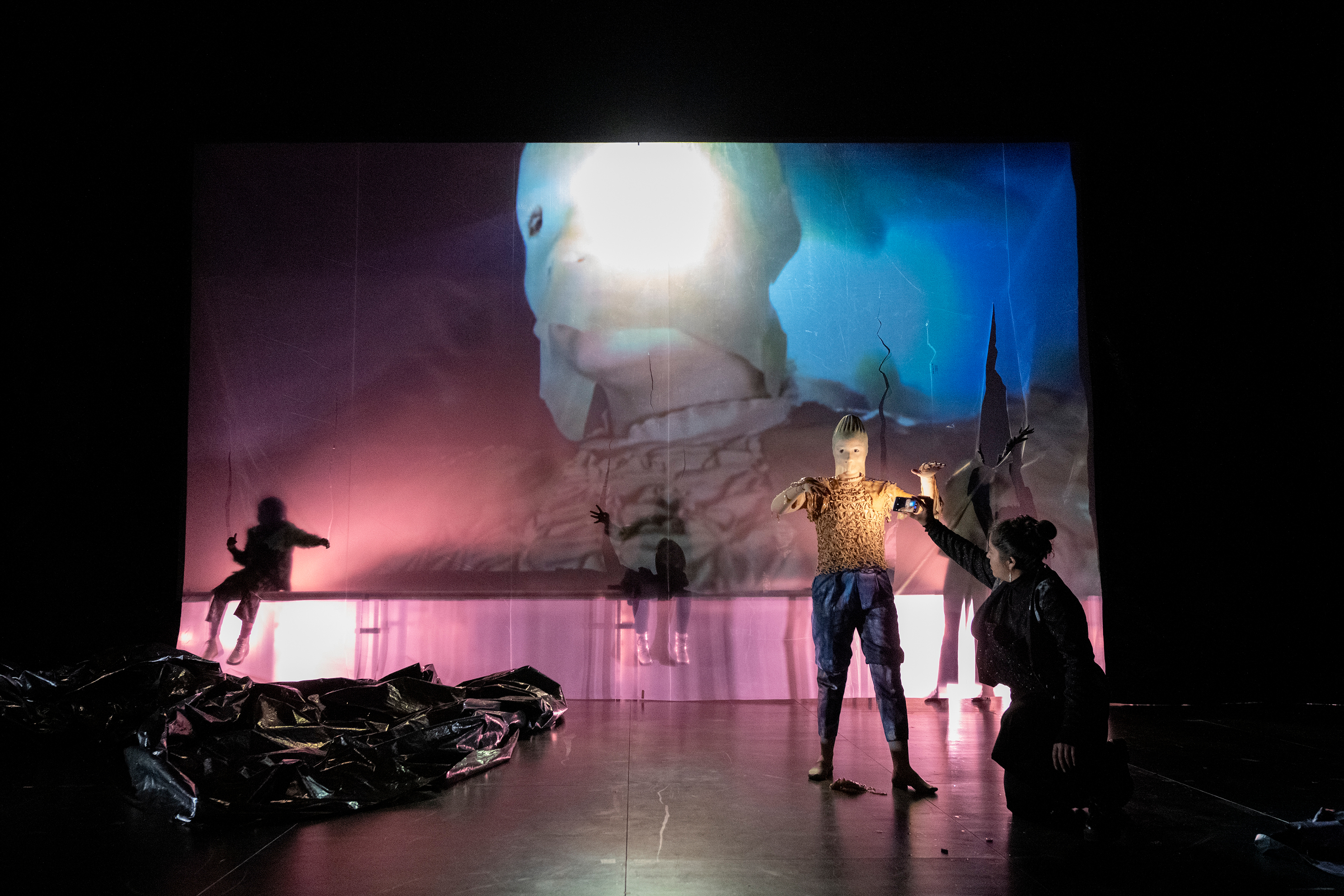 The performer moves across the stage with a kombucha mass in her arms, wearing a latex mask, abstract hand-painted pants, and a blouse made of elastic in a medium beige tone.